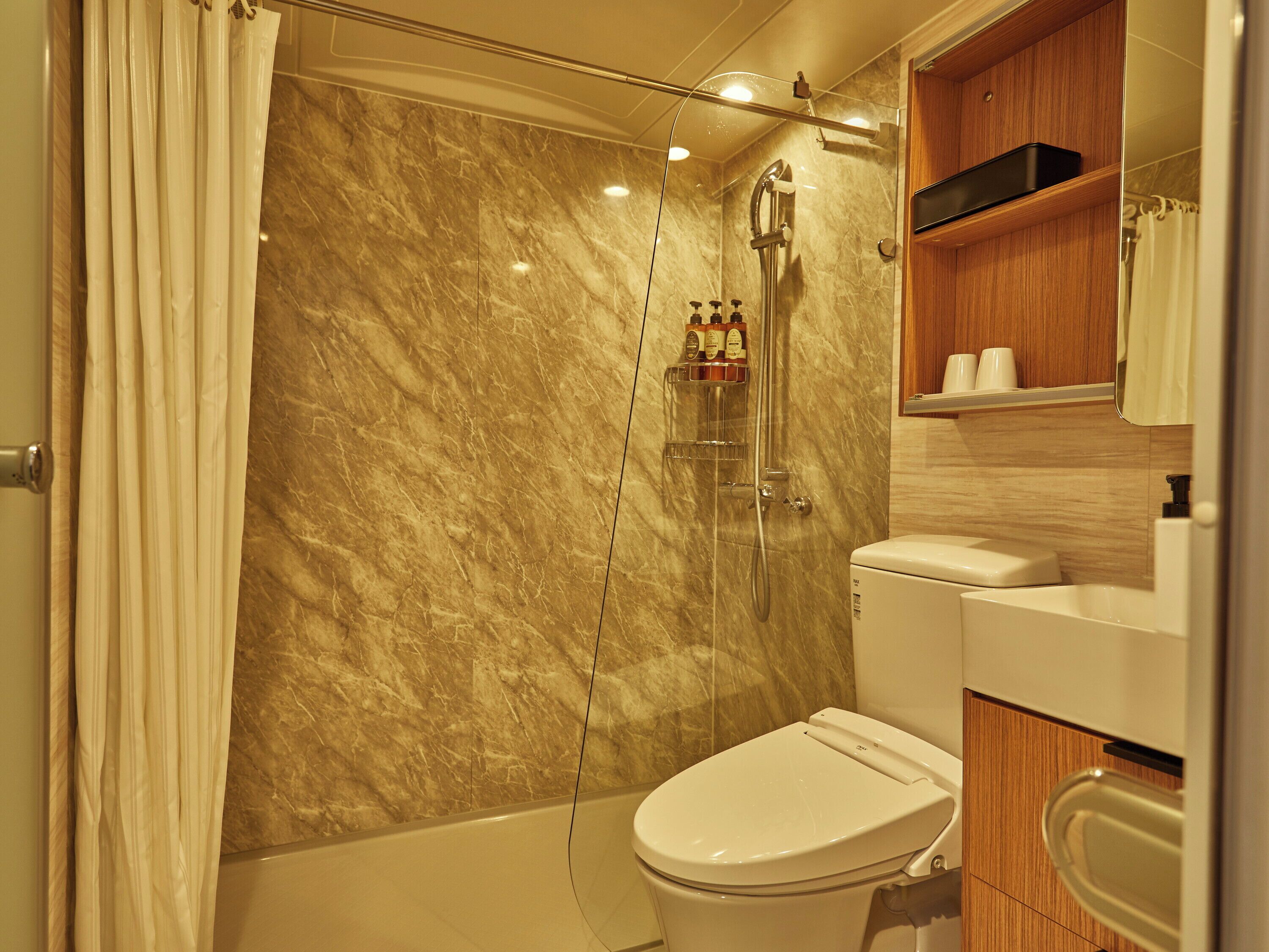 Un baño con pared de mármol, ducha, inodoro y repisa de madera con artículos de aseo.