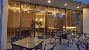 An outdoor seating area with tables and chairs, a glass wall separating the dining area from the interior, and a building with a modern design. - EZ HOTEL KIX Seaside (Izumisano)