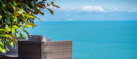 A wicker lounge chair overlooking a clear blue sea with distant land and a wooden deck.