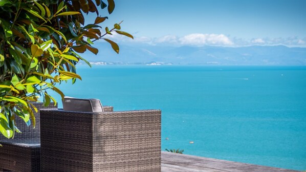 A wicker lounge chair overlooking a clear blue sea with distant land and a wooden deck.