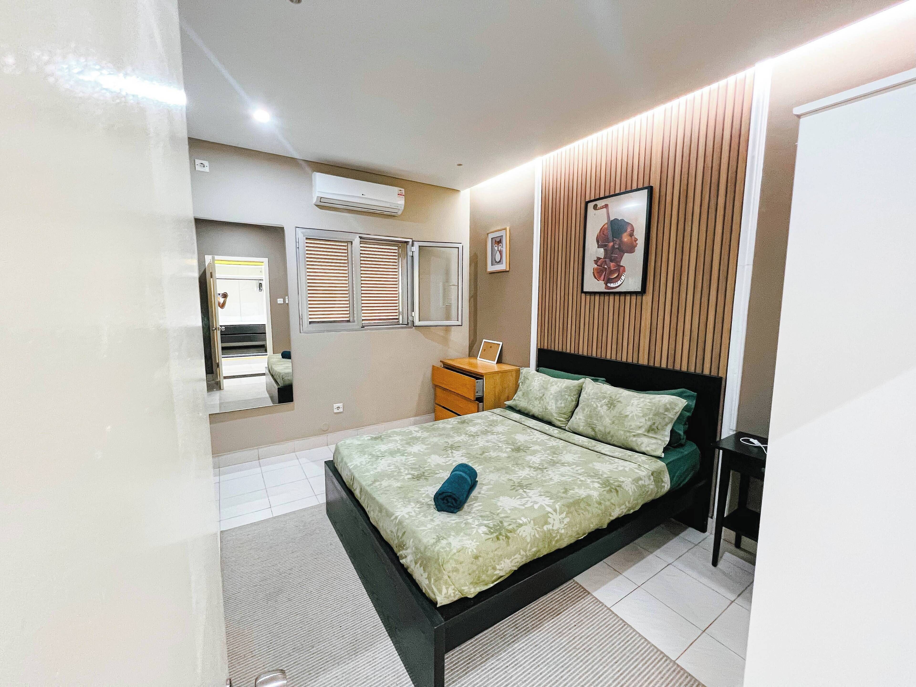 A bedroom with a bed, a bedside table, a mirror, an air conditioner, and a wall-mounted picture.