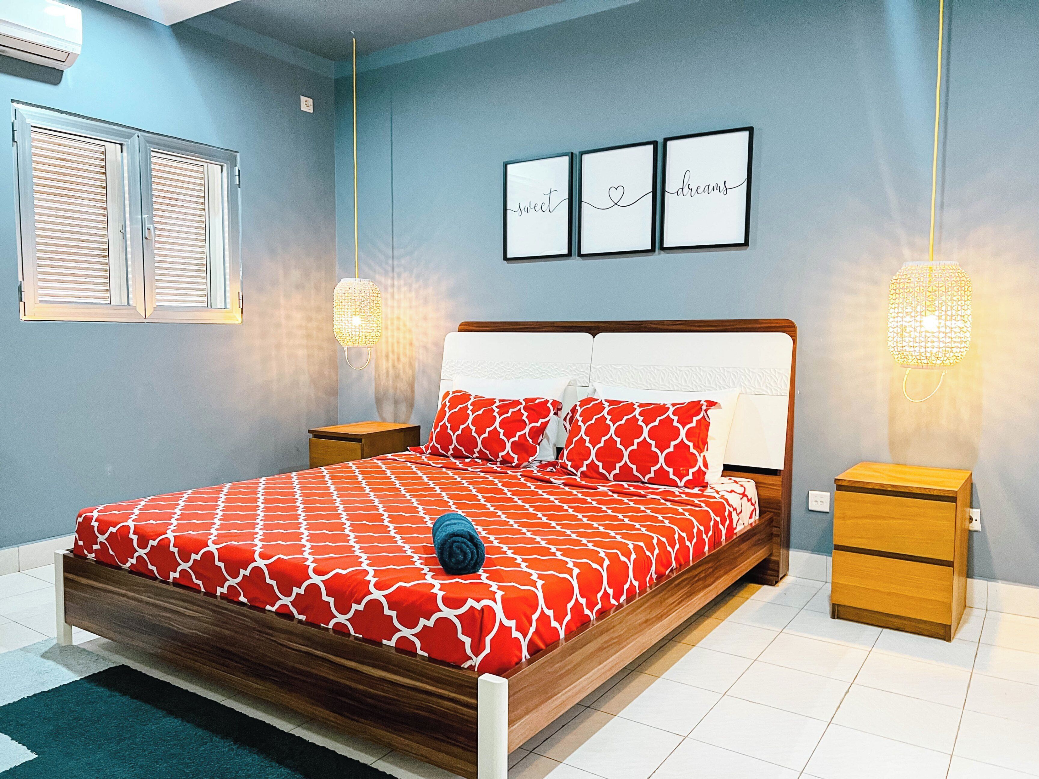 Cama bem arrumada com colcha estampada em vermelho e branco, duas mesas de cabeceira com abajures, e uma parede com três quadros emoldurados.