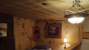 A wooden cabin room with a bed, a small table, a lamp, a painting, and a ceiling fan. - Our Guest house is a cozy get away, in a small town.  (Grey Eagle)