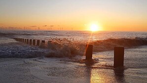 Solnedgang over en strand med bølger og vindmøller i horisonten.