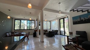 A spacious living area with a dining table, chairs, and a sofa. There are large windows providing natural light and views of greenery outside. - Casa Valle de Bravo la Peña  (Valle de Bravo)