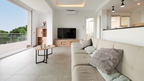 A modern living room with a large flat-screen TV, a sofa, a coffee table, and a view of greenery outside. - 2FIR02 Villa with private swimming pool (Lloret de Mar)