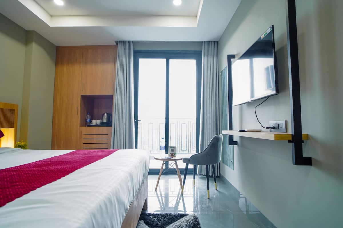 A hotel room with a bed, a desk, a chair, a TV mounted on the wall, and a balcony with curtains.