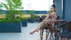 A person seated at a round table with a glass of drink, surrounded by wicker chairs and potted plants in an outdoor setting. - Saltstayz Select - Golf Course Road & Sector 57 (Gurugram)