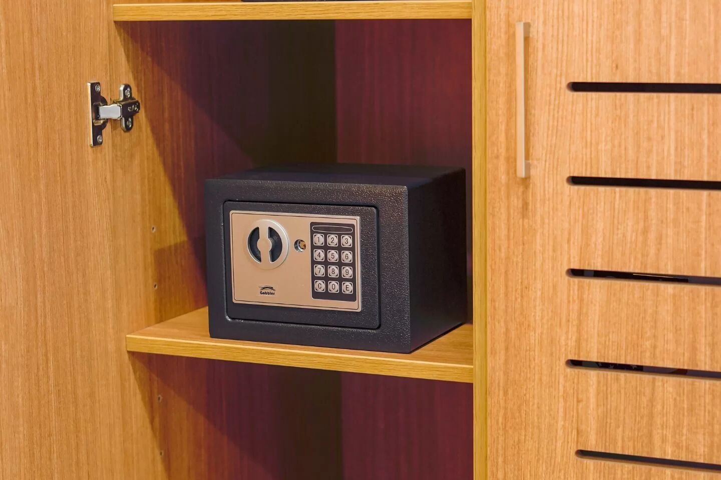 A safe with a keypad lock on a wooden shelf inside a wooden cabinet.