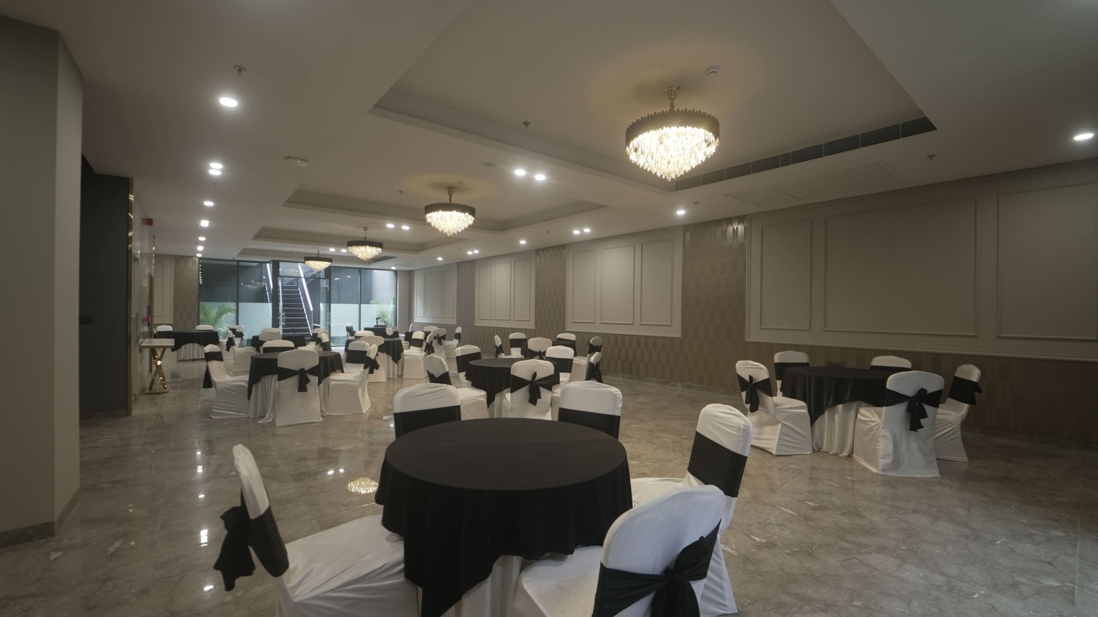 A spacious banquet hall with round tables, chairs with black sashes, and chandeliers.