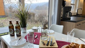 A dining table set with plates, cutlery, glasses, and a bottle of beer, with a view of mountains through the window.