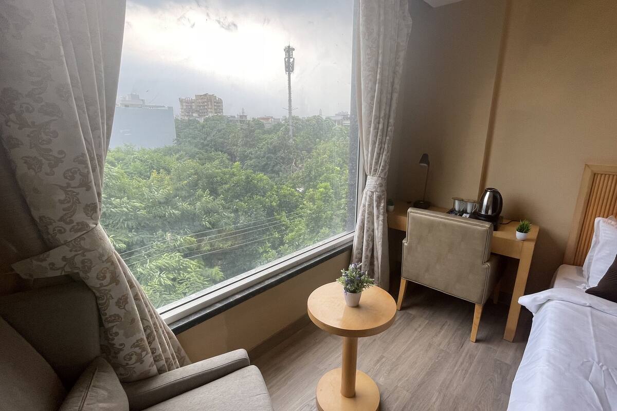 A hotel room with a bed, a small round table with a plant, a nightstand with a kettle, and a view of a cityscape through the window.