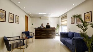 A hotel reception area with a reception desk, two receptionists, a blue sofa, a blue armchair, and framed artwork on the walls. - Saltstayz Select - Galleria Market & Golf Course Road (Gurugram)
