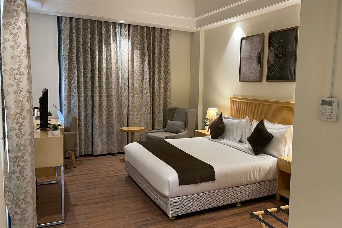 A hotel room with a large bed, a desk with a computer, a chair, a lamp, and a window with curtains.