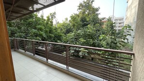 A balcony with a metal railing overlooking a garden with trees and a building in the background. - Saltstayz Select - Galleria Market & Golf Course Road (Gurugram)