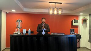 A reception desk with a man standing behind it, a coffee machine, a bottle of hand sanitizer, and a sign with QR code. - Saltstayz Express - Golf Course Road & DLF Phase-1 (Gurugram)