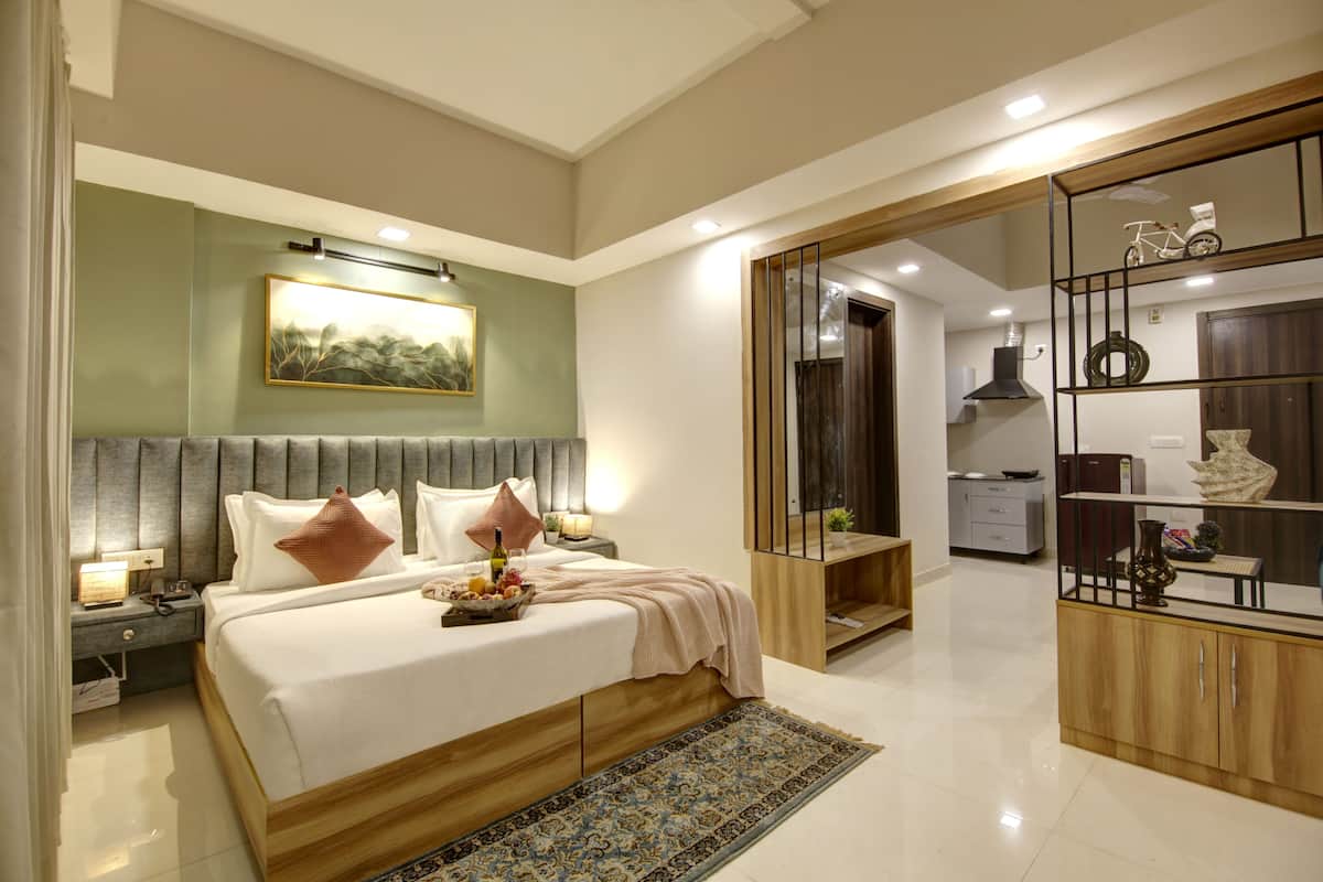 A modern bedroom with a large bed, bedside tables, a nightstand, a wooden wardrobe, and a small kitchen area.
