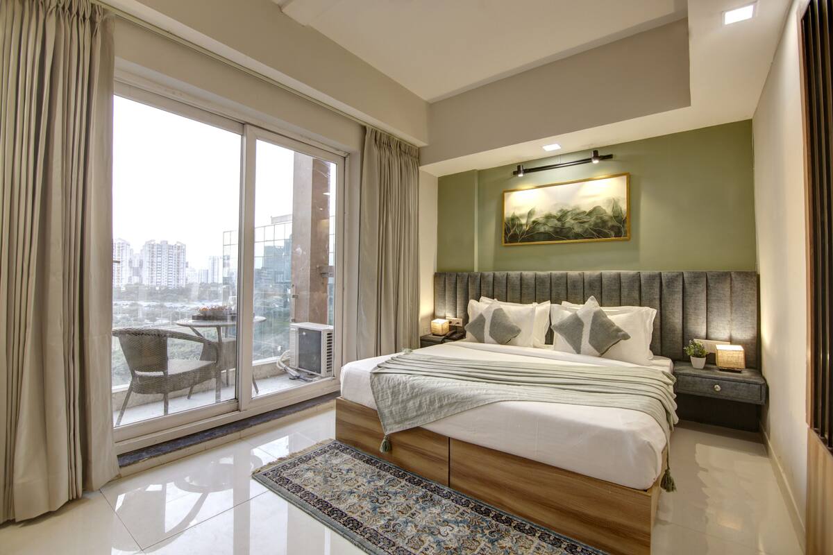 A modern bedroom with a large bed, a city view through floor-to-ceiling windows, a dining area with a table and chairs, and a small balcony.