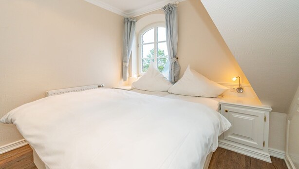 A bedroom with a large bed, a window with curtains, a bedside table with a lamp, and a radiator. - Atlantis - NEU ab 2024 in Unserer Vermittlung! (Sylt-Ost)