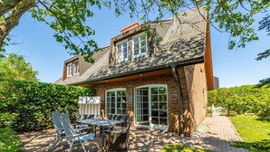 A two-story house with a brick exterior, large windows, and a patio area with outdoor furniture. - Atlantis - NEU ab 2024 in Unserer Vermittlung! (Sylt-Ost)