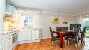 A modern kitchen with white cabinetry, a dining area with a wooden table and chairs, and a large window providing natural light. - Atlantis - NEU ab 2024 in Unserer Vermittlung! (Sylt-Ost)
