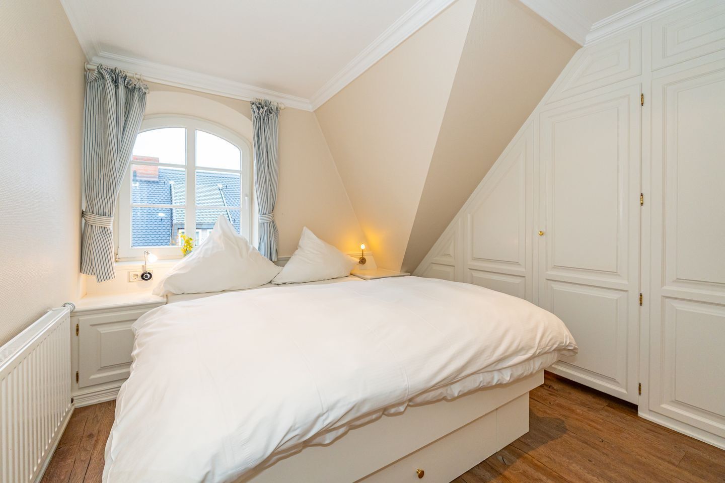 A bedroom with a large bed, a window with curtains, a nightstand with a lamp, and a wardrobe.