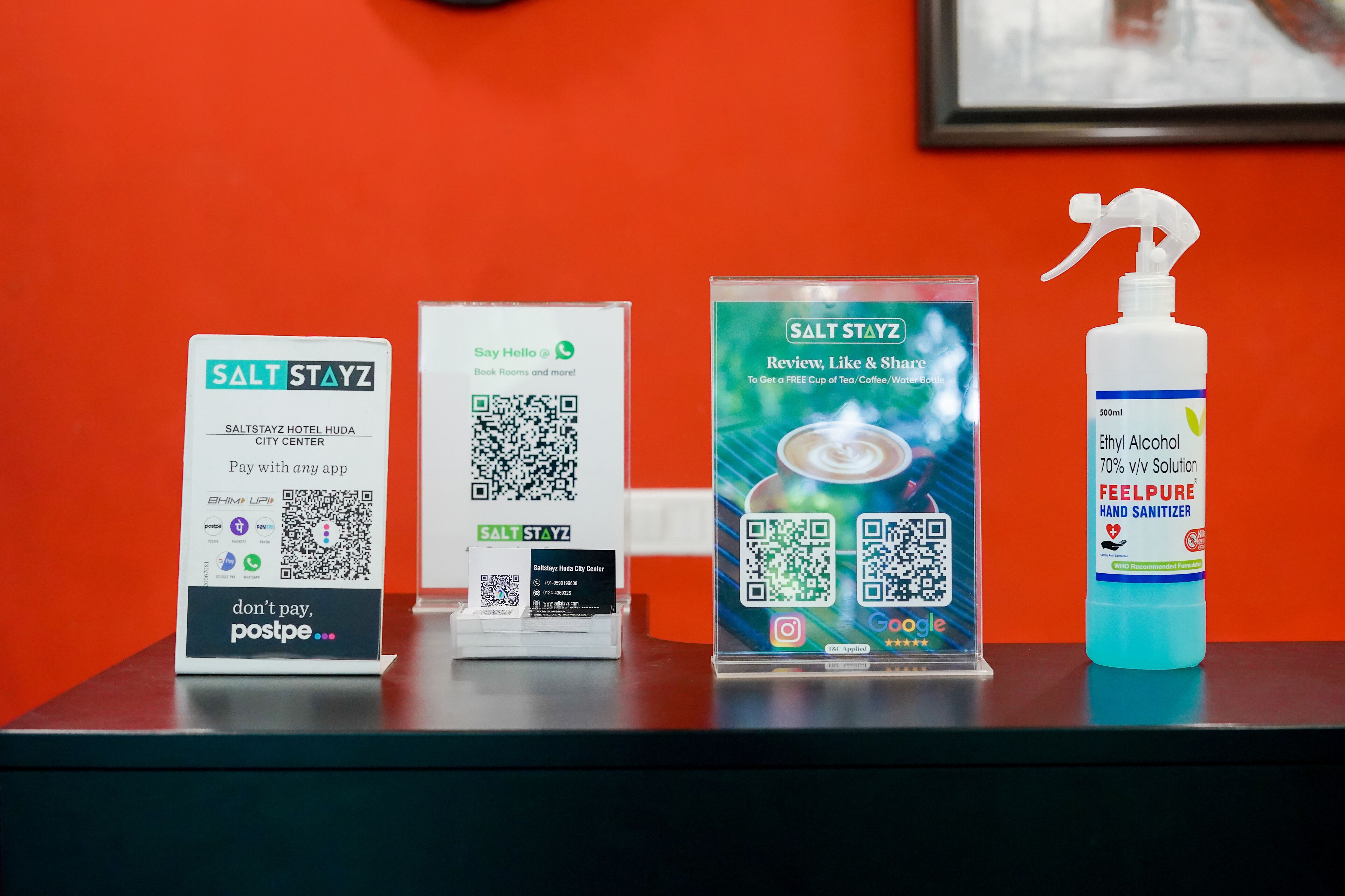 A hotel reception desk with a sign for SALT STAYZ HOTEL HUDA CITY CENTER, a 'Stay Hello' sign, a promotional poster for SALT STAYZ, and a hand sanitizer bottle.
