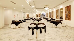 Un salón de banquetes con varias mesas redondas, cada una con sillas blancas y cubrecubiertos negros, y una araña colgante.