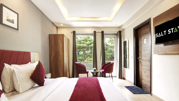 Habitación de hotel con una cama grande, un corredor rojo, dos sillas, un televisor y un balcón con cortinas.