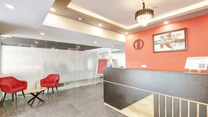 Zona de recepción con diseño moderno, que incluye un mostrador de recepción, dos sillones rojos, un separador de vidrio y una araña.
