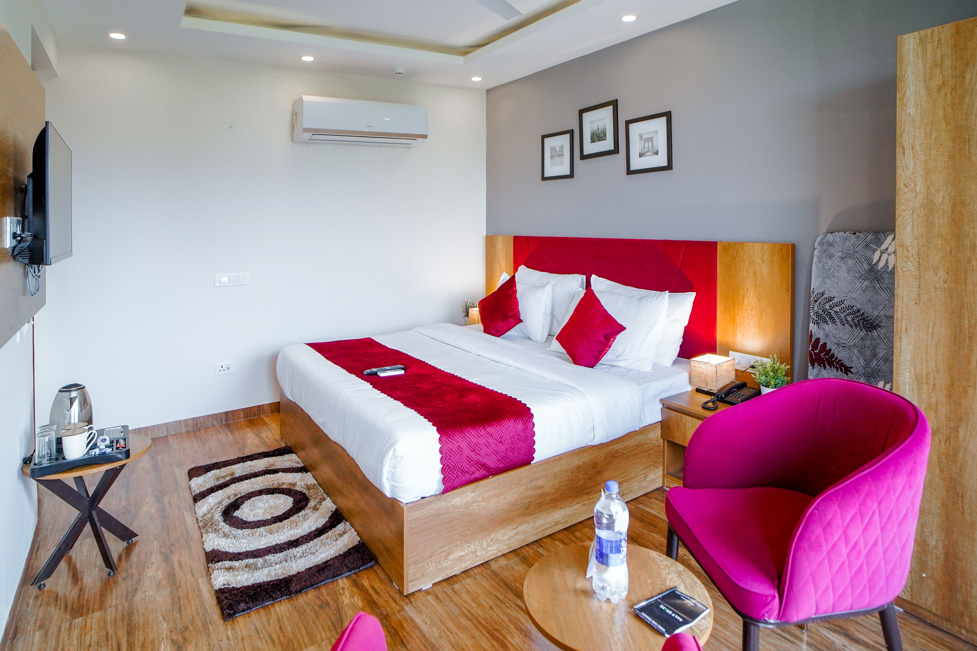 A modern hotel room with a wooden bed, red and white bedding, a red chair, a small table with a water bottle, and a wall-mounted TV.