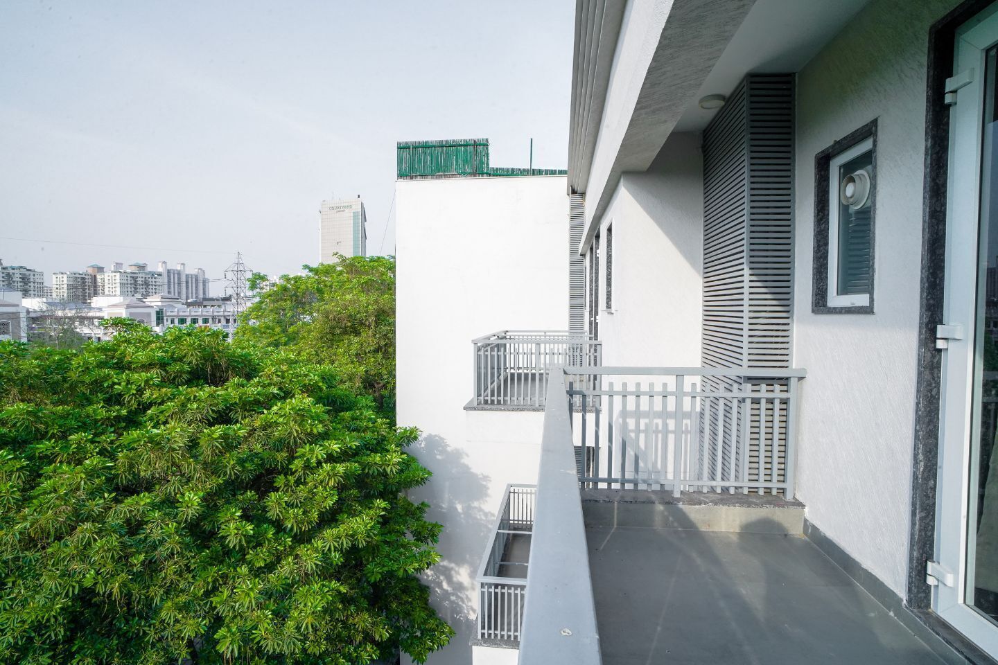 A balcony with a white railing, a small window with shutters, and a view of a cityscape with buildings in the background.