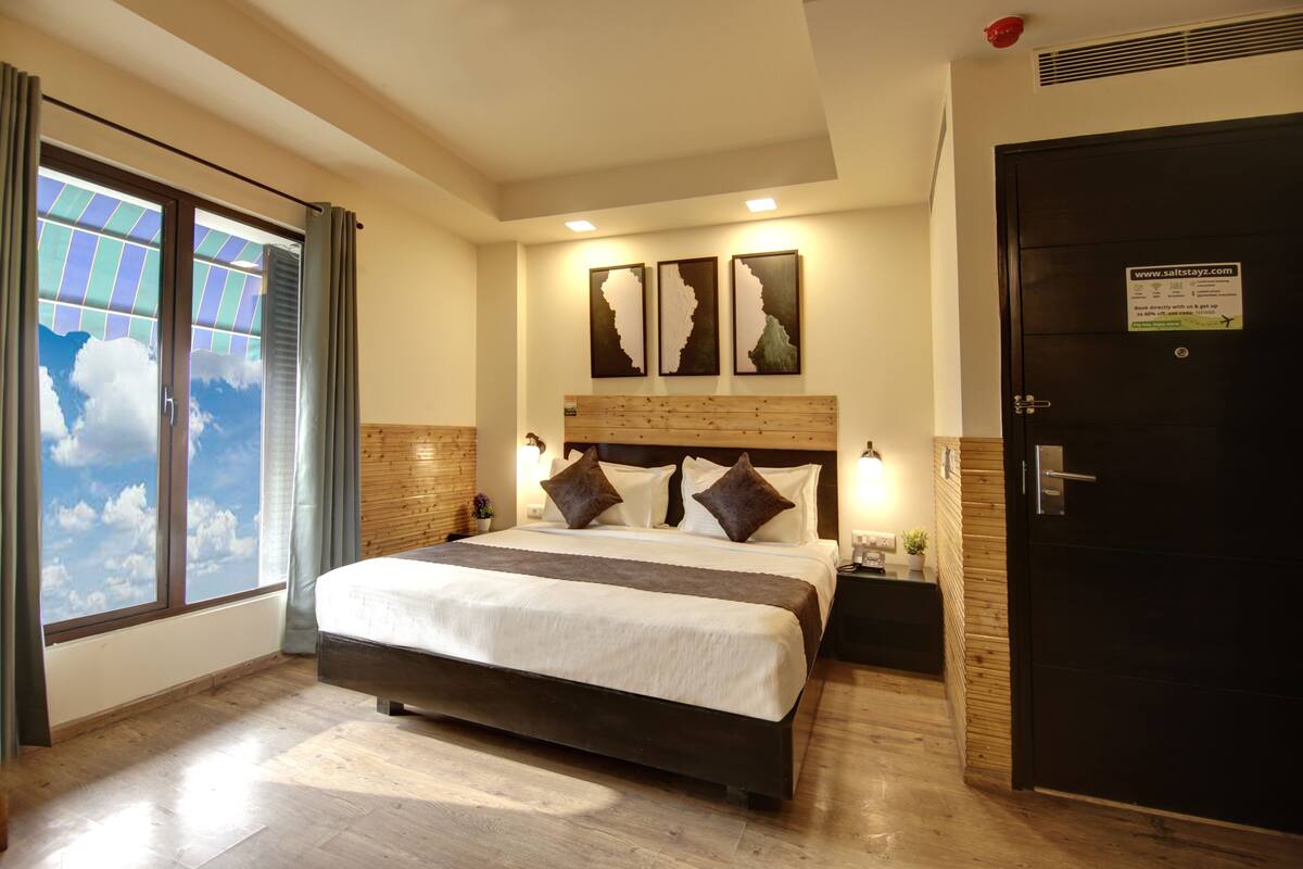 Una habitación de hotel con una cama grande, un cabecero de madera, una mesita de noche, una puerta negra y vistas al cielo a través de la ventana.