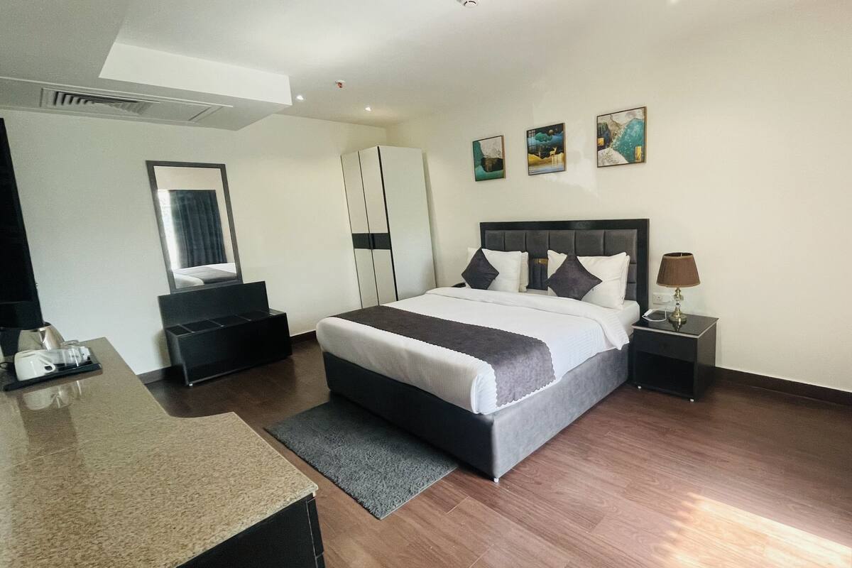 A modern hotel room with a large bed, a desk, a chair, a nightstand with a lamp, and artwork on the walls.