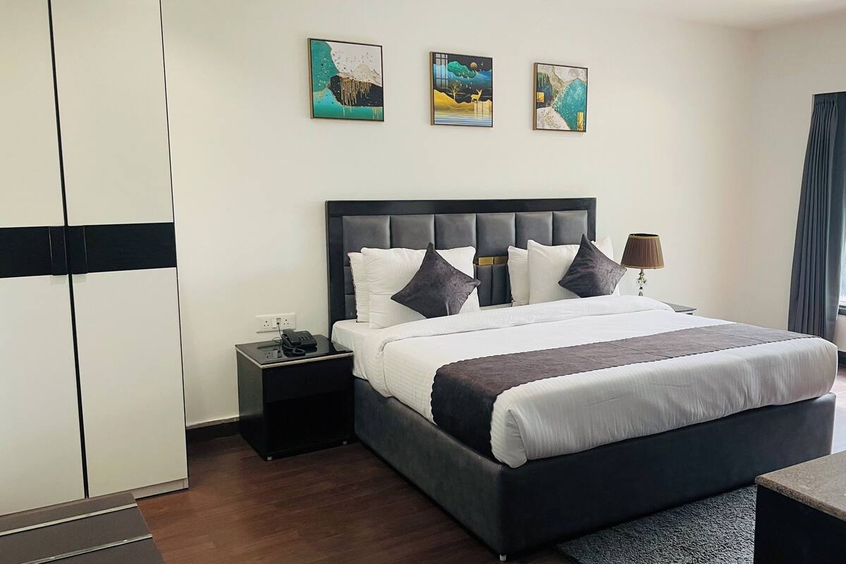 A modern hotel room with a large bed, a nightstand, a desk, and a wardrobe. There are three paintings on the wall and a window with curtains.