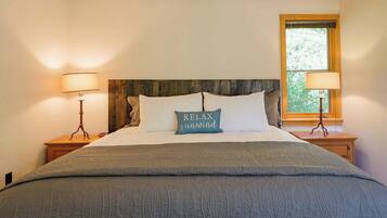Una cama bien hecha con una almohada que dice 'RELAX & unwind', dos lámparas de noche y una ventana con vista a árboles.