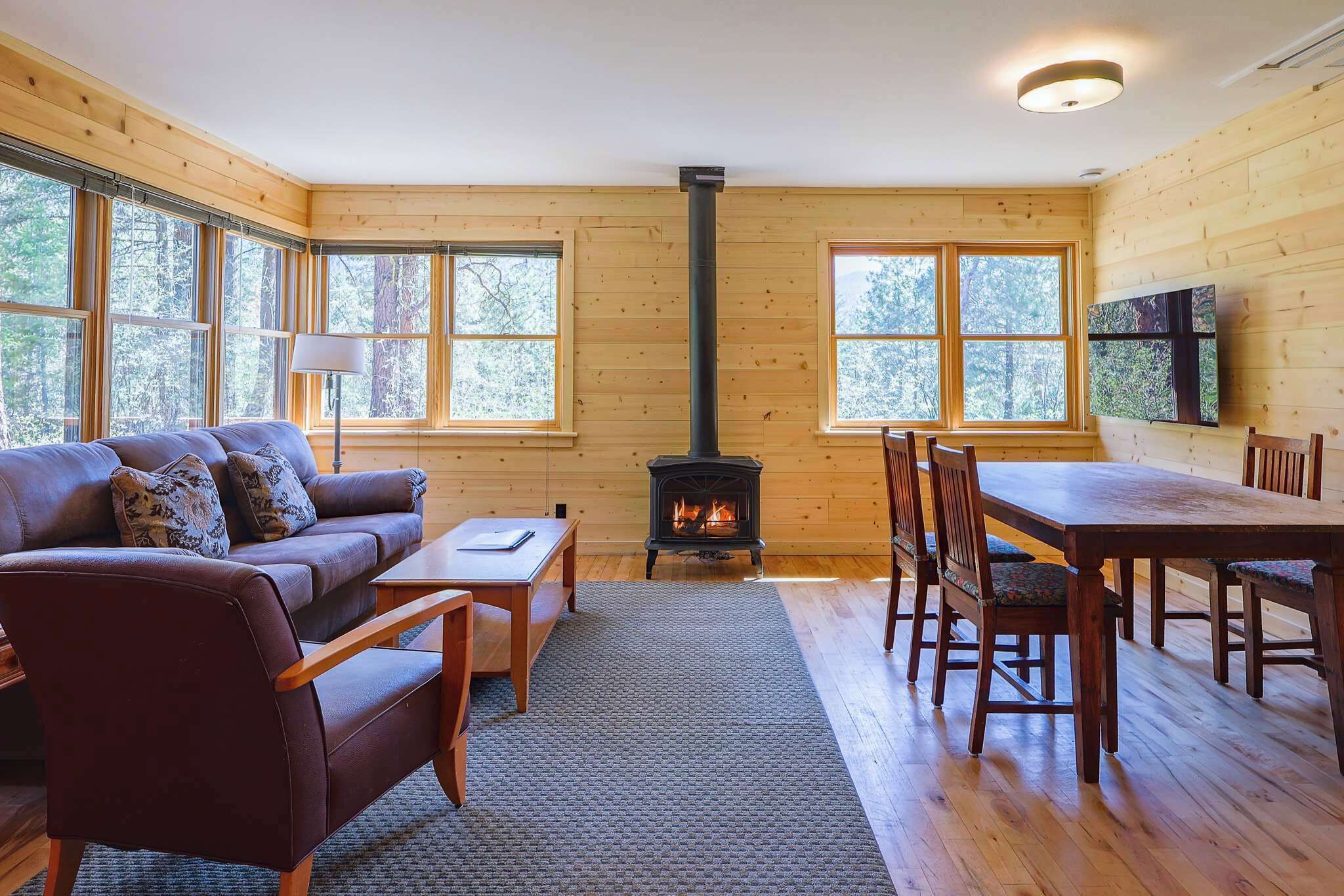 Un interior de cabaña acogedor con un sofá, una mesa de comedor, sillas, una chimenea y ventanas grandes.