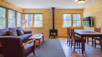 Un interior de cabaña acogedor con un sofá, una mesa de comedor, sillas, una chimenea y ventanas grandes.