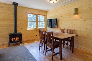 A wooden interior with a fireplace, a dining table with chairs, and a window with a view of trees.