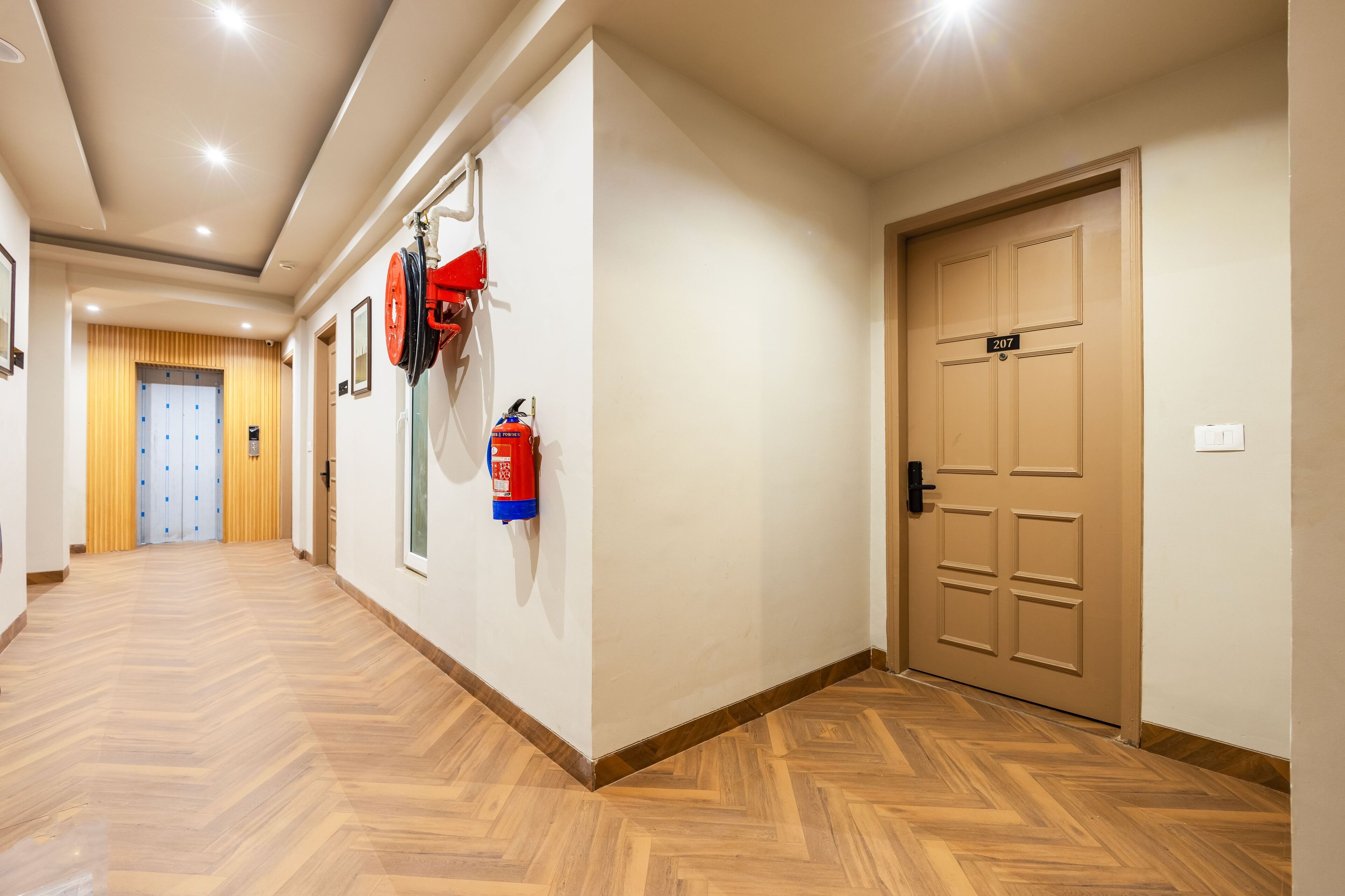Un pasillo con suelo de madera, un extintor y una puerta con el número 207.
