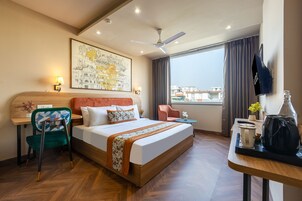 Habitación de hotel con cama, escritorio y silla, televisor, ventana con cortinas y vista panorámica de la ciudad.