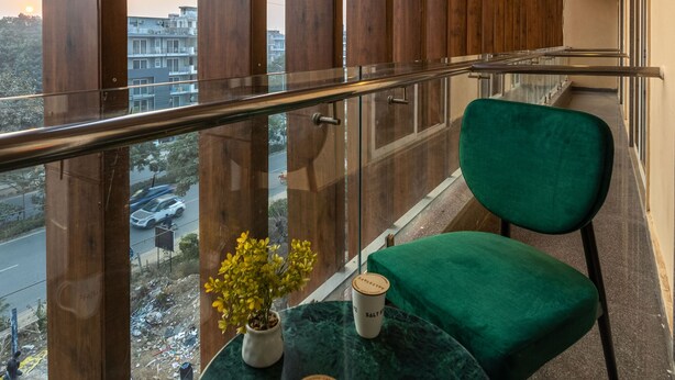 A balcony with a green cushioned chair, a small table with a potted plant and two coffee cups, and a view of a street and buildings. - Saltstayz Select- Medicity Near Medanta Hospital (Gurugram)