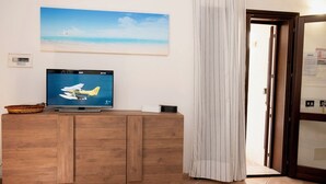 Huoneessa on puinen kaappi, televisio, jossa näkyy lentokone, rantamaisemaa esittävä taulu ja verholla varustettu ovi.