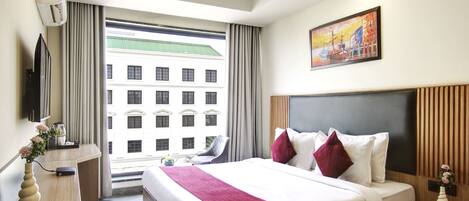 A modern hotel room with a large bed, a desk, a chair, a TV, and a view of a building outside.
