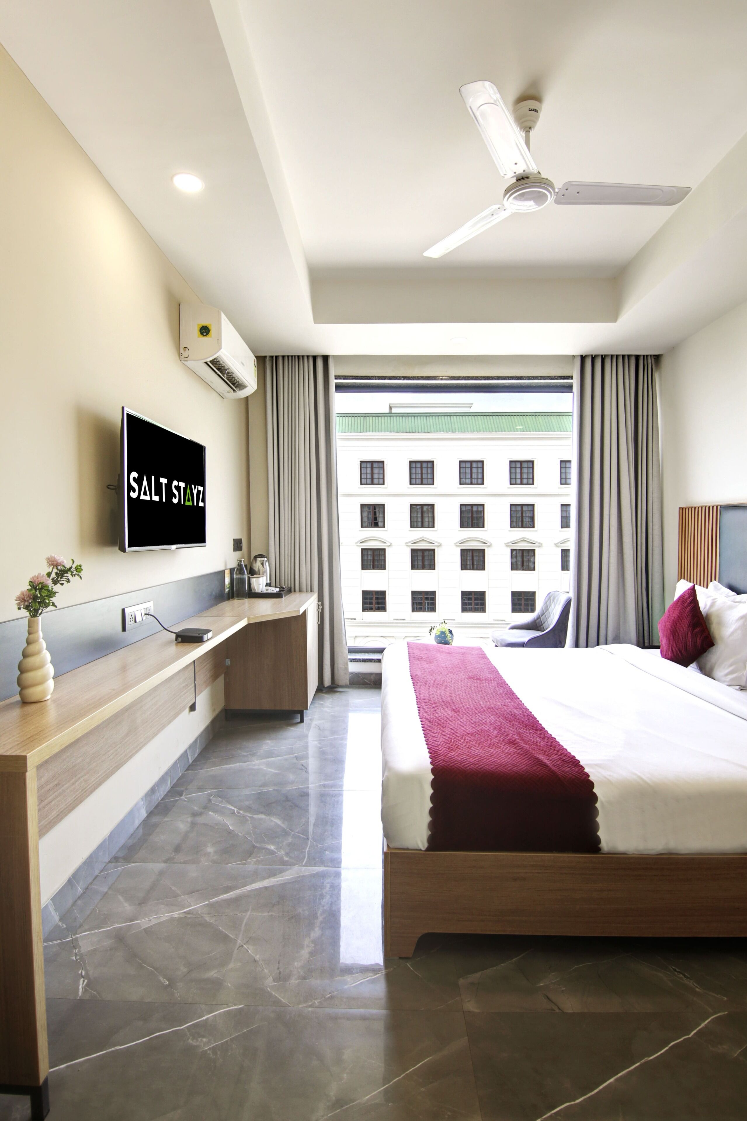 A modern hotel room with a bed, a desk, a television, and a view of a building outside.