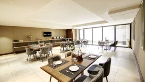 A modern hotel dining area with multiple tables and chairs, a kitchen in the background, and large windows providing natural light. - Saltstayz Premier – Cyber Hub (Gurugram)