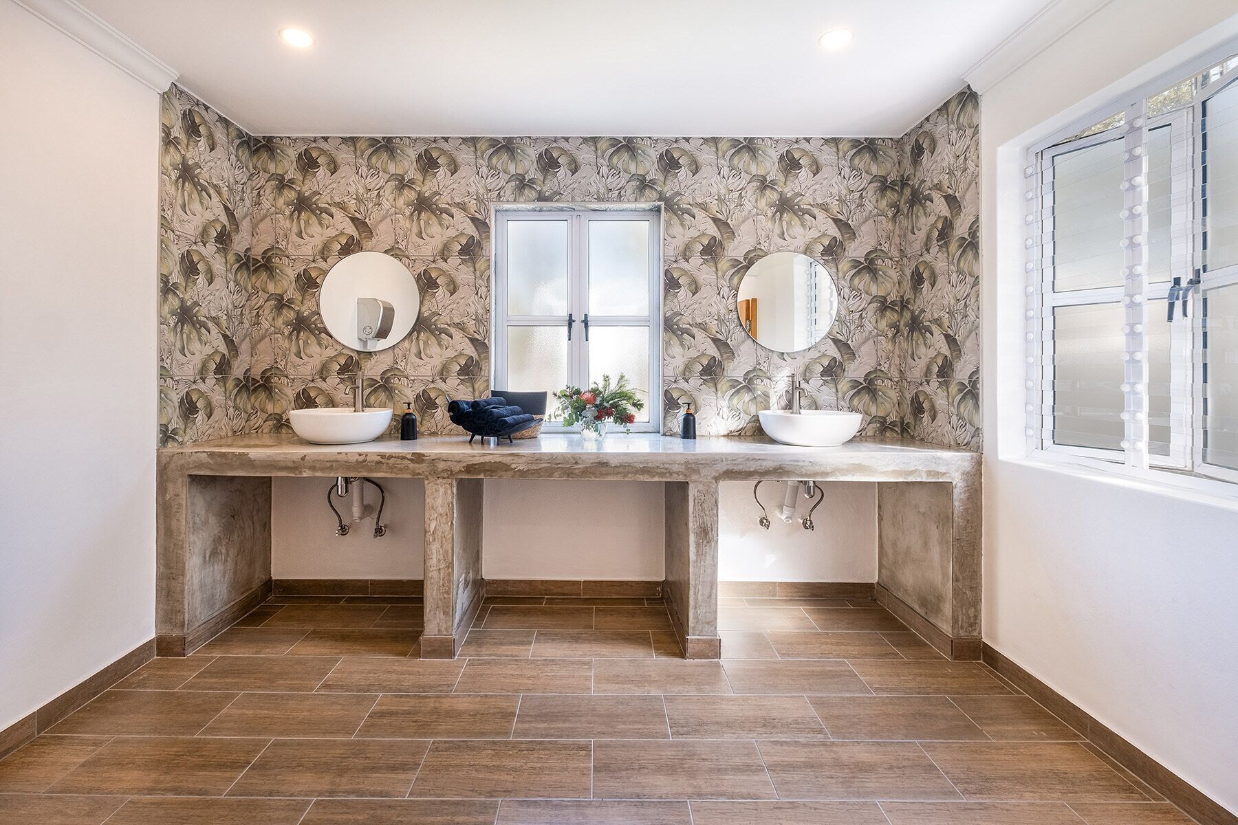 Une salle de bain avec deux lavabos, chacun agrémenté d’un miroir rond, d’un lavabo sur colonne blanc et d’un flacon de savon. Les murs sont recouverts d’un papier peint à motifs floraux, et une fenêtre avec des stores se trouve sur le côté droit.