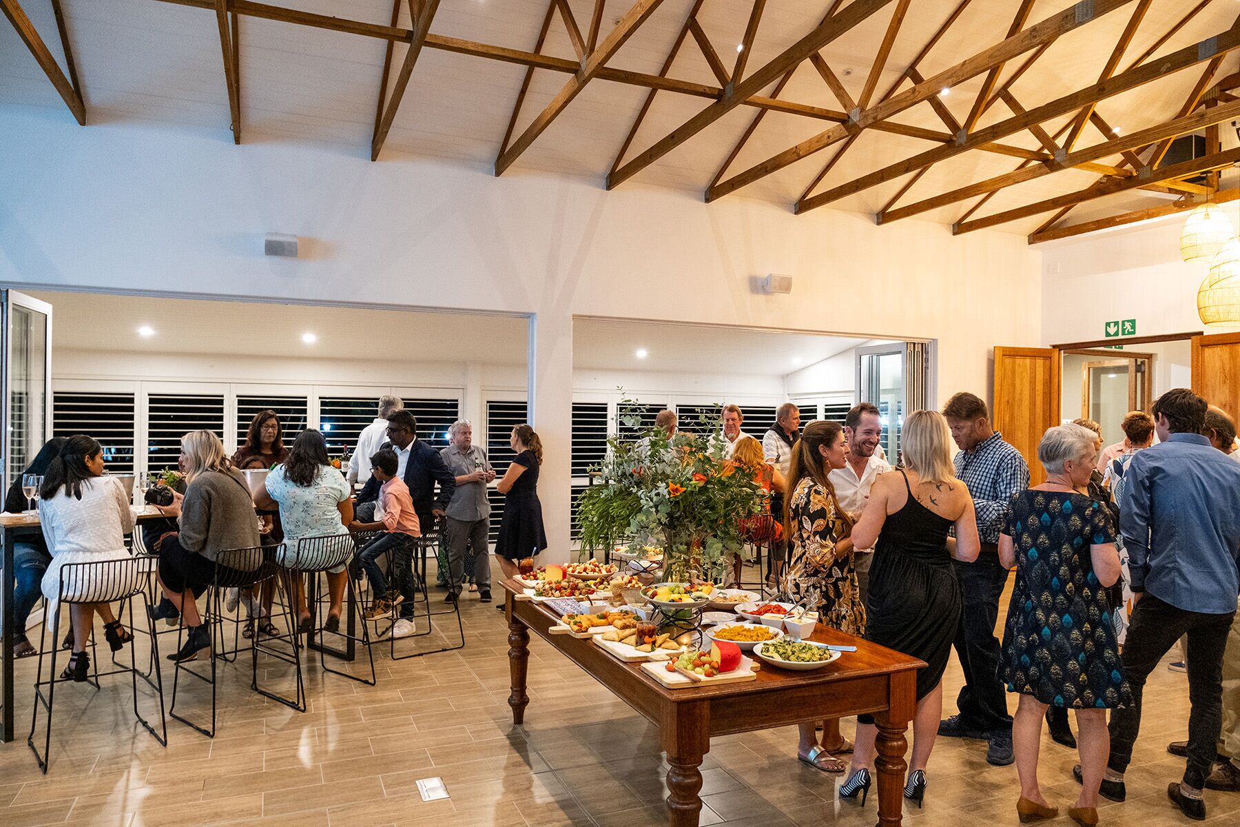 A spacious indoor event with a high ceiling and wooden beams, featuring a long table with various food items and a group of people gathered around.
