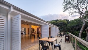 Une terrasse en bois avec une table et des chaises, un coin cuisine visible à travers des portes ouvertes, et une vue sur les arbres et la verdure.