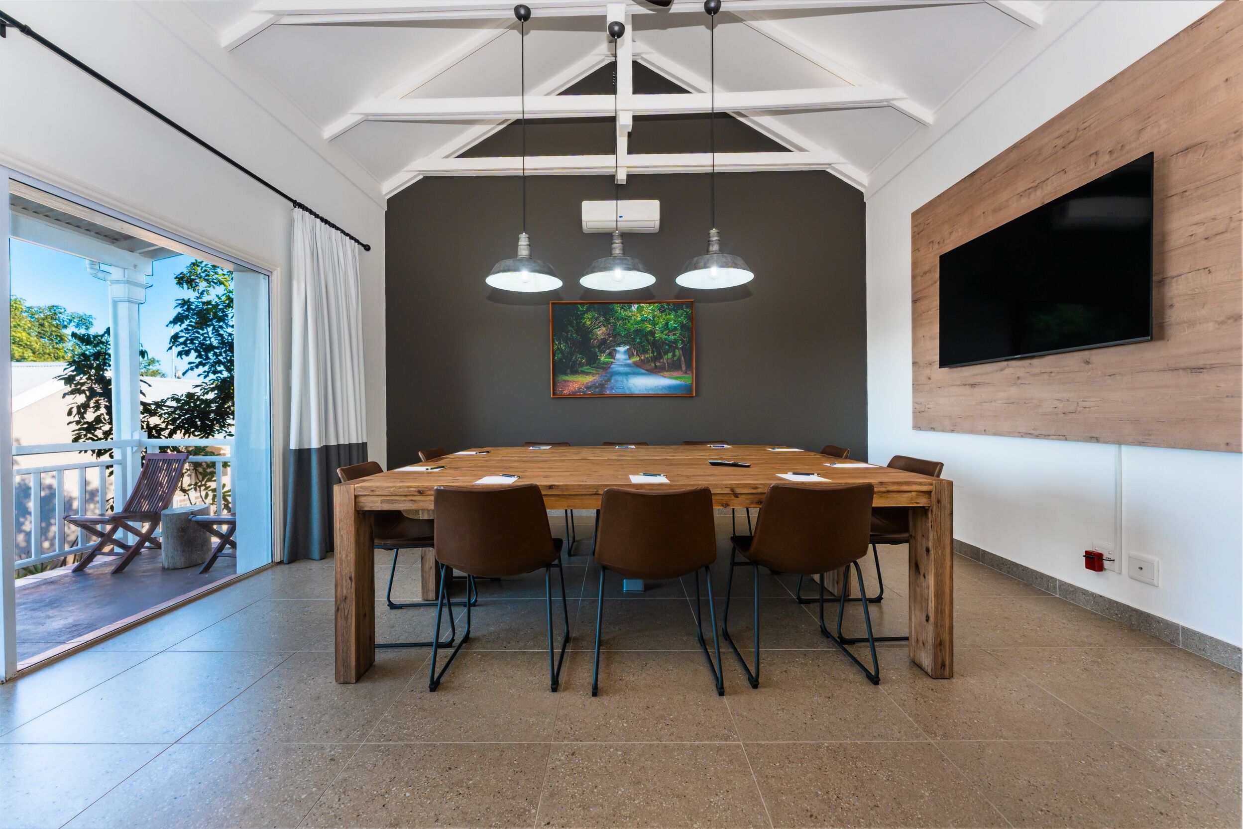 Une salle de conférence avec une grande table en bois, quatre chaises, des luminaires suspendus, un tableau et un téléviseur fixé au mur.